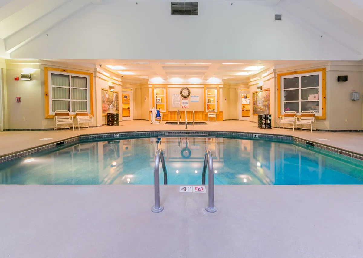 A bright, welcoming indoor pool area with comfortable poolside seating, tasteful wall decor, and a glimpse of the reception area. The calm water and soft lighting create an inviting space for both relaxation and socializing, suitable for swimmers and those looking to unwind alike.