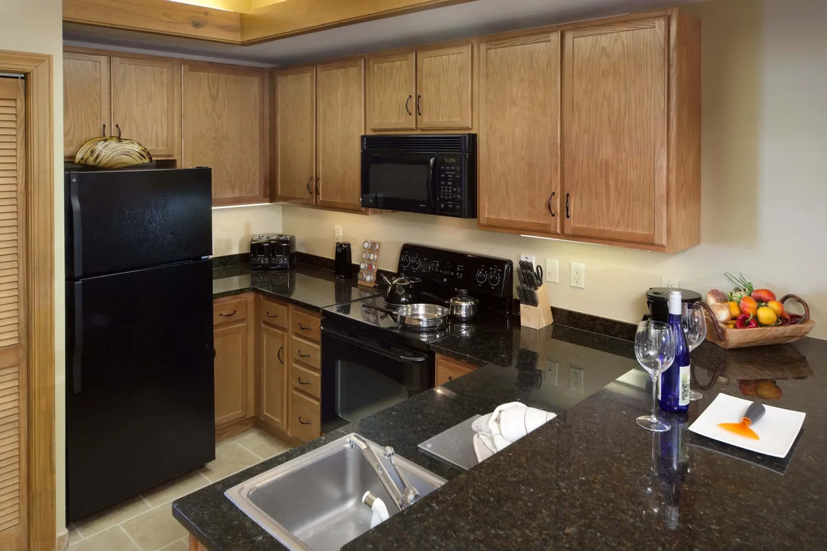 This image showcases a fully-equipped kitchen with a modern black refrigerator, granite countertops, and wooden cabinets. A fruit bowl adds a welcoming touch, and the space is clearly designed for cooking, entertaining, and family meals.