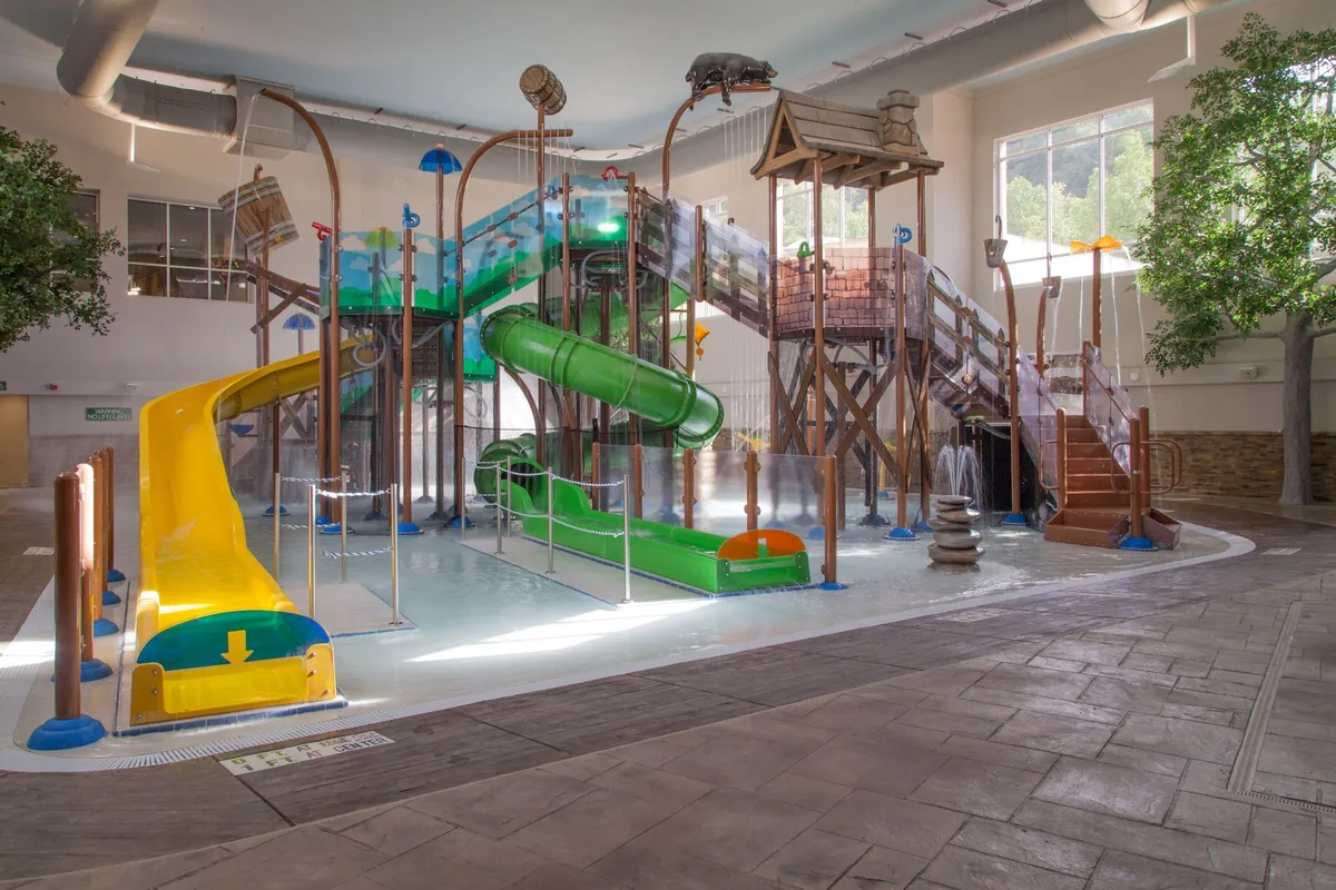 This lively interior image captures a bustling indoor water park complete with colorful water slides, a splash area, and an interactive play structure surrounded by greenery. Children and families can be seen enjoying various water-based activities, radiating joy and excitement.