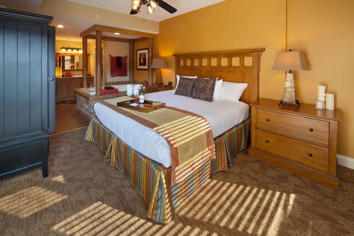 A spacious primary bedroom featuring a plush king-sized bed dressed in decorative bedding. The wooden headboard and matching nightstands with lamps create a warm, inviting atmosphere. Sunlight filters softly through the windows, and an open space leads seamlessly to the adjacent bathroom, emphasizing comfort and convenience.