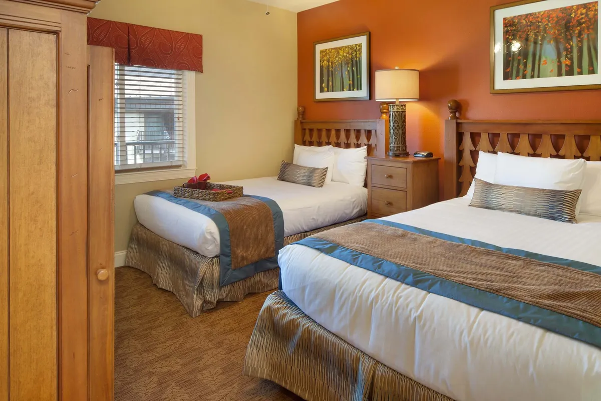 A cozy second bedroom furnished with two double beds, wooden furniture, a decorative lamp, and artwork on the wall. The room exudes warmth and is naturally lit, inviting families or groups to enjoy bedtime stories, morning coffee in bed, or simply unwind together.