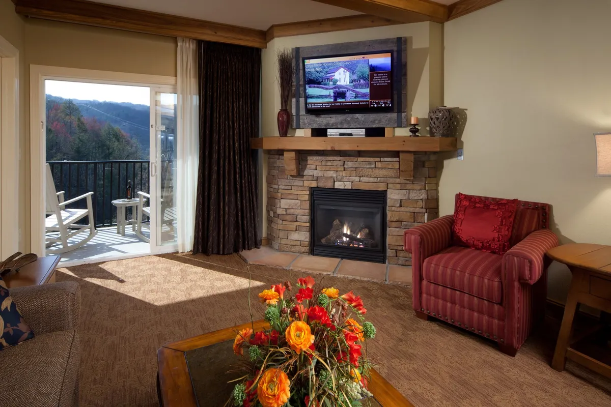 The image features a cozy, stylish living area with a glowing fireplace, plush seating, a flat-screen TV, and a vibrant flower arrangement. Sunlight pours in through large windows, offering a glimpse of the scenic Smoky Mountains, inviting guests to relax together and soak in the tranquil mountain atmosphere.